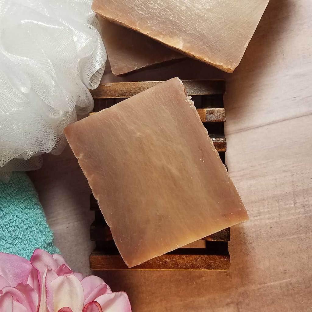 Handmade coconut soap bar on a wooden soap dish surrounded by a loofah, towel, and flower, showcasing a natural bath experience.