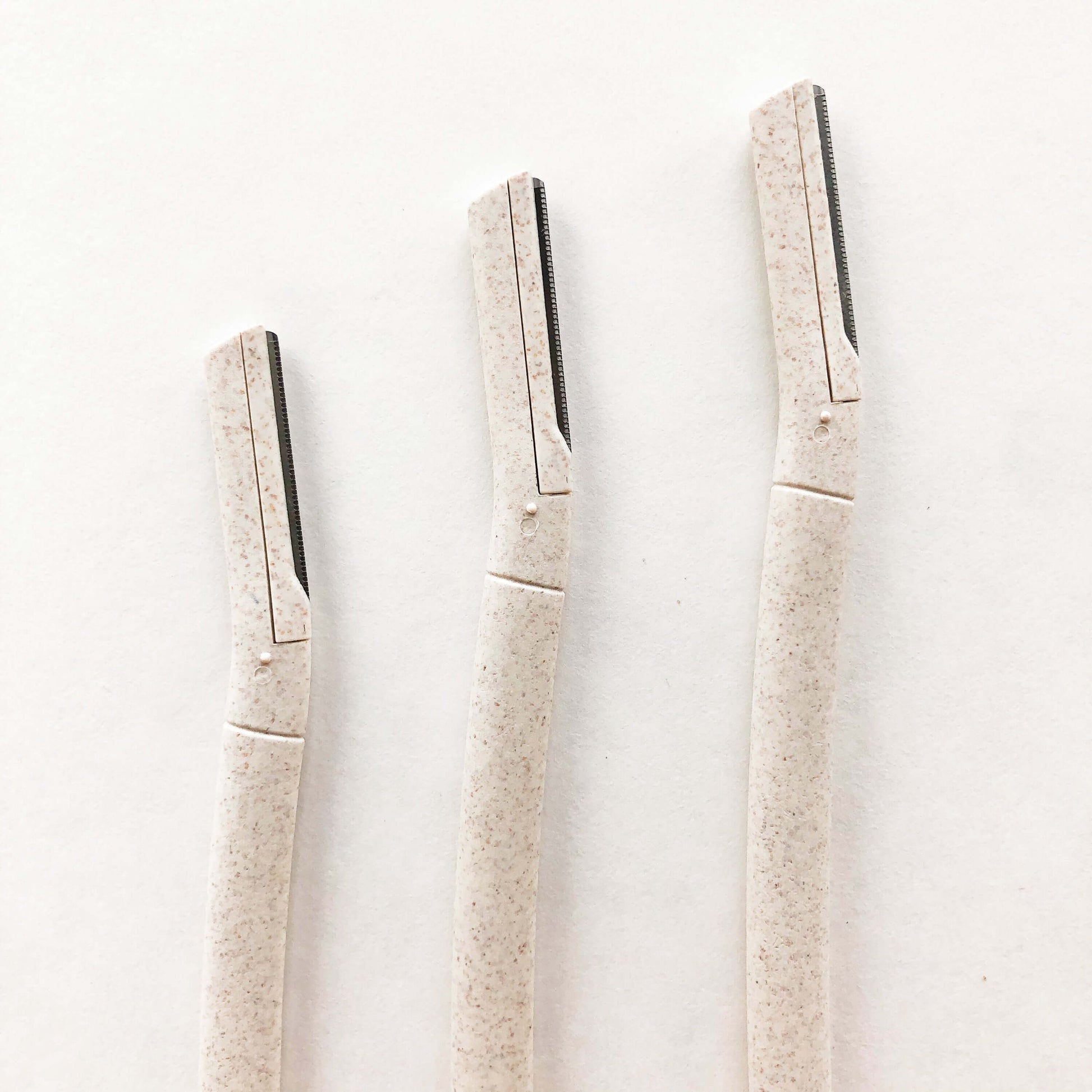 Three biodegradable wheat straw eyebrow razors lined up on a plain background, eco-friendly beauty tools for flawless brows.