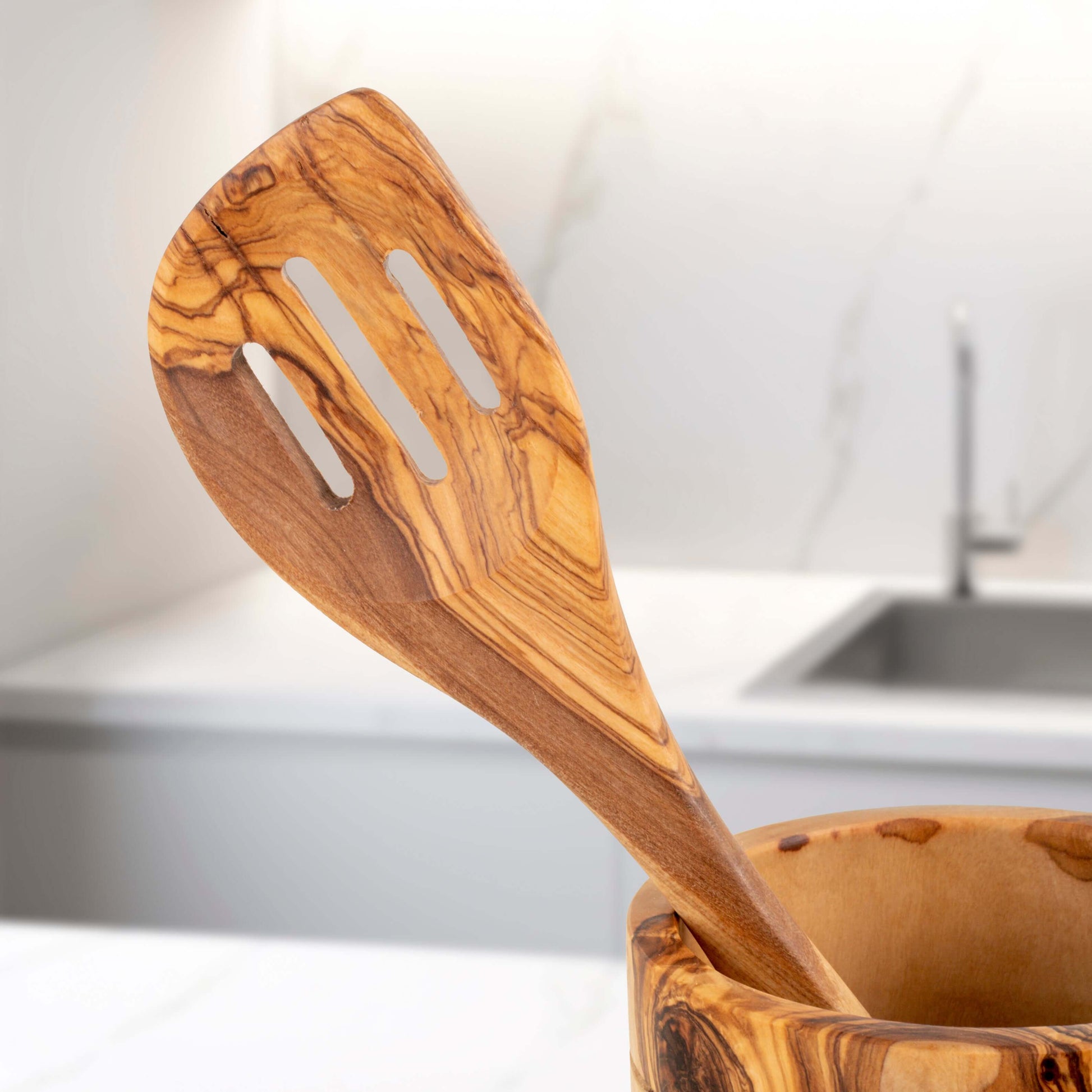Handmade olive wood slotted spatula resting in a wooden holder in a modern kitchen.