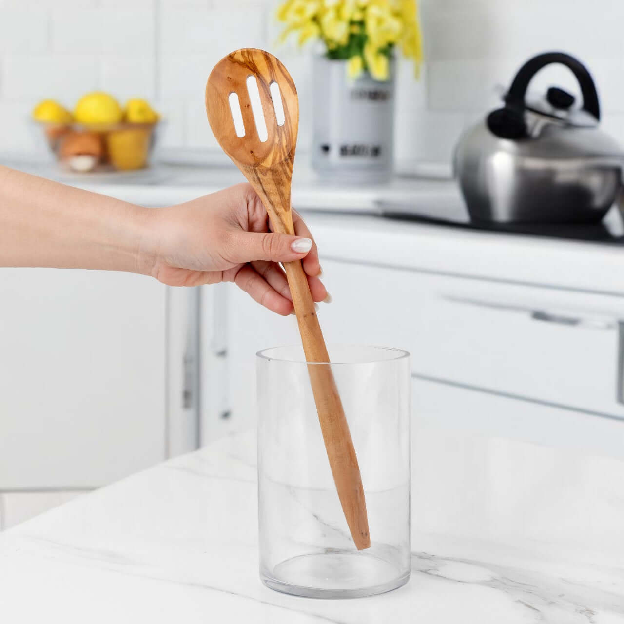 Hand holding a 35cm olive wood slotted spoon above a glass container in a modern kitchen setting.