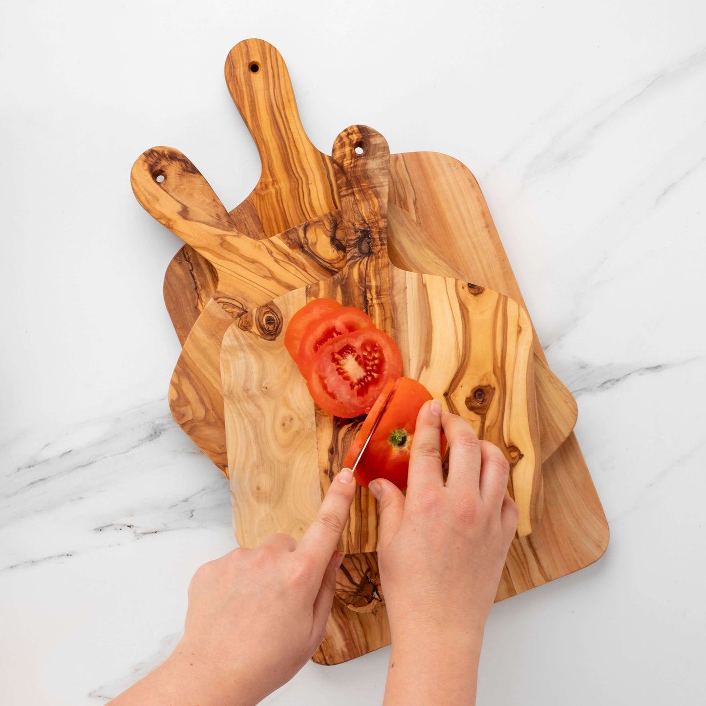 Person slicing a tomato on olive wood cutting boards with handles, showcasing their unique grain patterns.