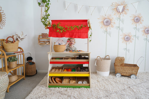 Child playing at a colorful wooden market stall surrounded by toys in a bright, cozy room.