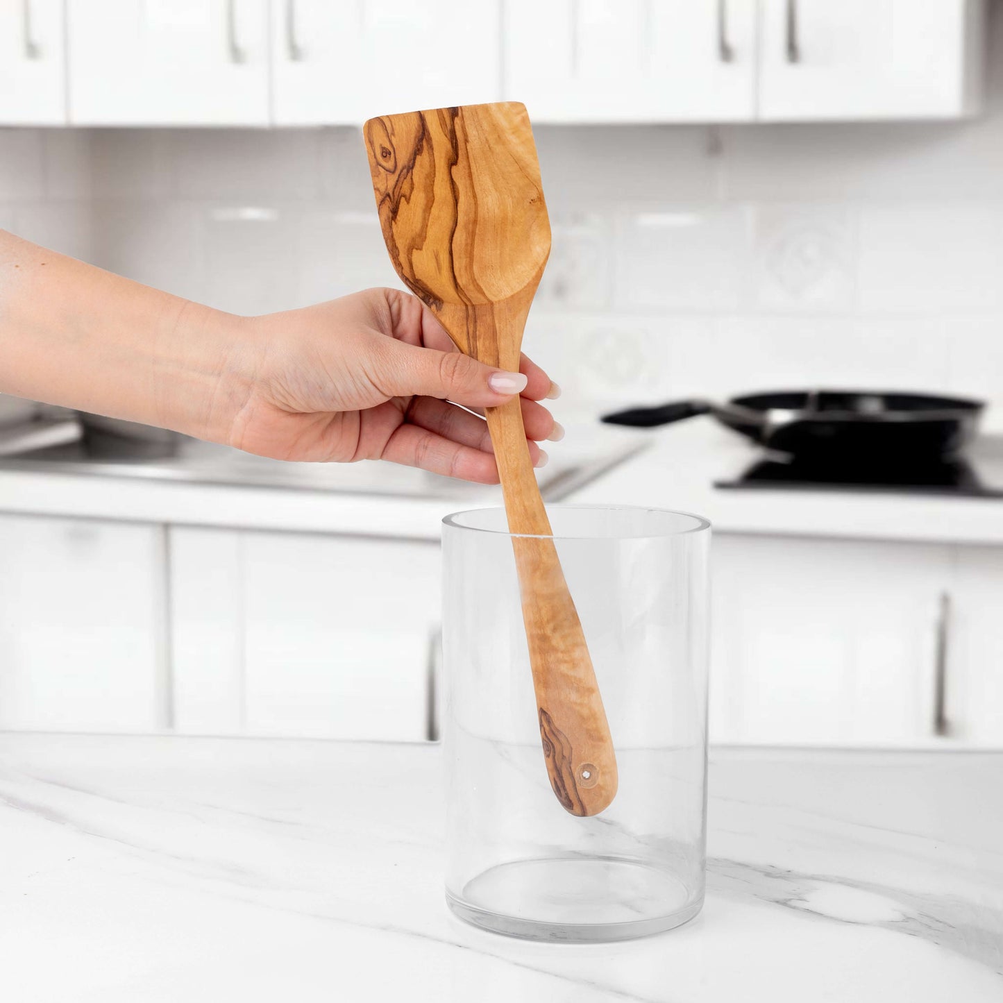 Handmade 11.8" olive wood spatula being held above a glass container in a modern kitchen.