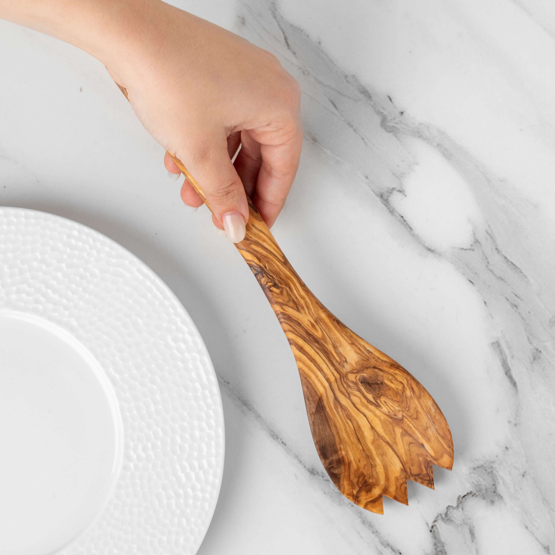 Handmade olive wood salad spork being held above a white plate on a marble surface.