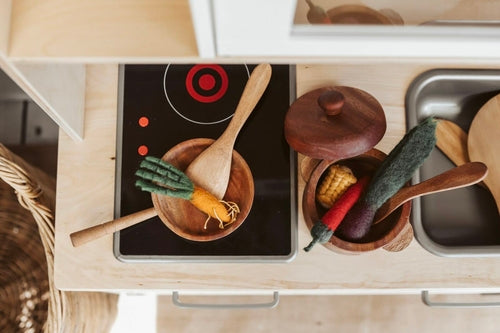Top view of wooden cooking utensils and felt vegetables in bowls on a toy kitchen countertop.